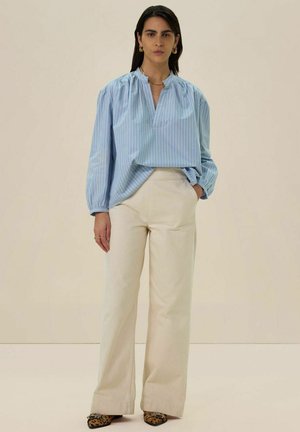 Blouse à rayures bleu clair avec une coupe ample, encolure froncée et manches longues, associée à un pantalon large crème et des chaussures à motifs.
