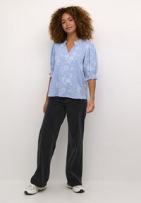 Lichtblauwe bloemenblouse met korte pofmouwen en een V-hals, gecombineerd met donkere broek met wijde pijpen en witte sneakers.