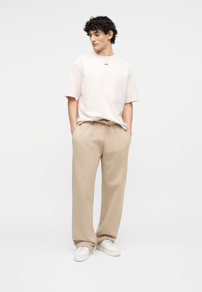 Νεαρός άνδρας στέκεται με τα χέρια στις τσέπες μπεζ παντελονιών με κορδόνια, φοράει άνετο λευκό t-shirt και λευκά αθλητικά παπούτσια ενάντια σε απλό φόντο.