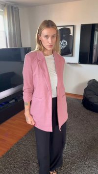 Femme blonde portant un blazer rose, une chemise blanche et un pantalon noir, debout dans un salon avec une télévision, des œuvres d'art encadrées et un tapis.