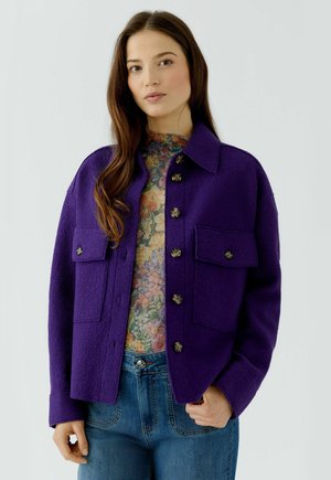 Junge Frau trägt eine violette Wolljacke mit Knöpfen über einem floralen Oberteil und blauen Jeans, steht vor einem einfarbigen hellen Hintergrund.