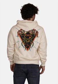 Felpa crema con una grafica di ragno su uno sfondo a forma di diamante in nero e arancione. Presenta un cappuccio con stringa e polsini a costine.