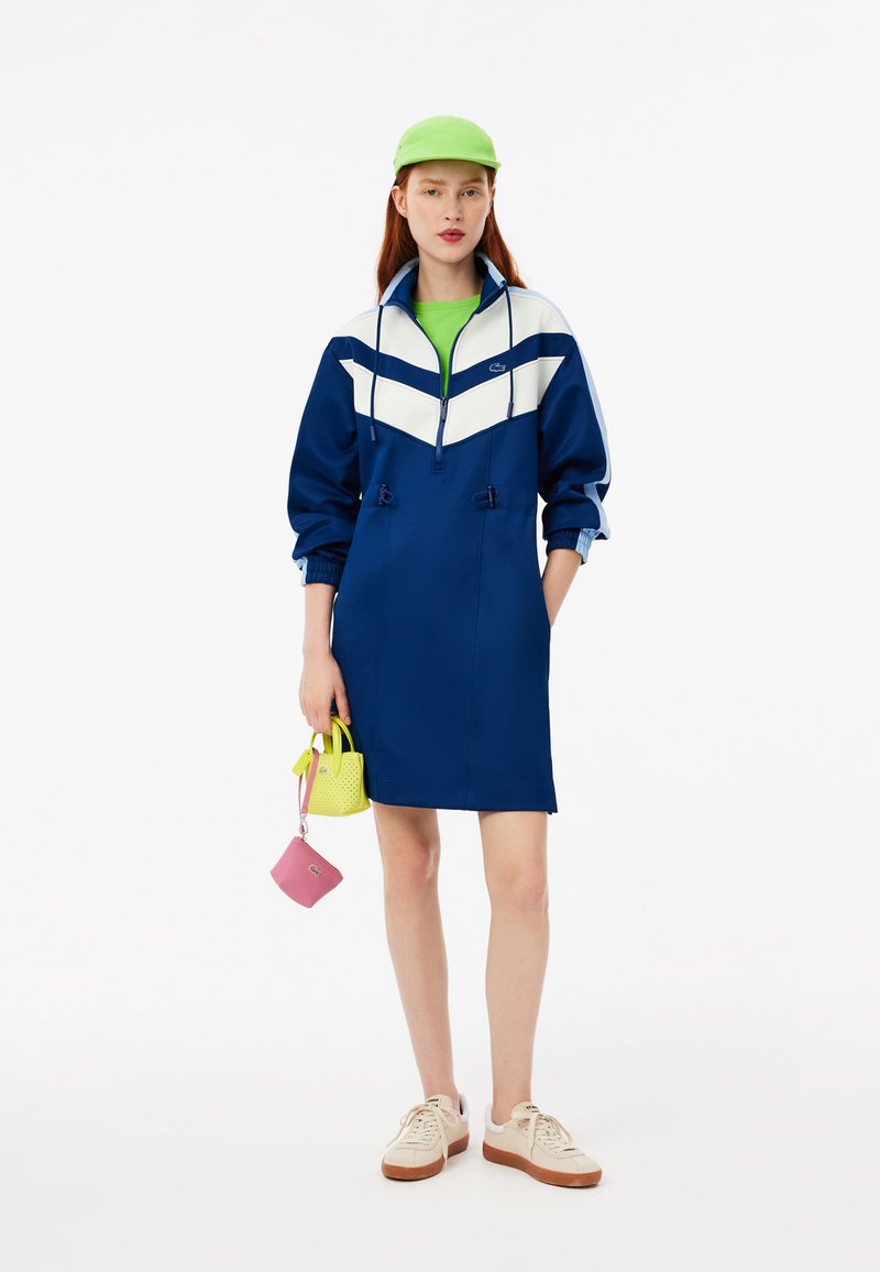 Vestido cortaviento azul marino con diseño de chevron blanco, puños elásticos, dos cordones y un bolsillo tipo canguro. Par de zapatillas blancas y bolsos.