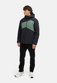 Svart och grön färgblockerad pufferjacka med dragkedja, huva och texturerad tyg, kombinerad med svarta byxor och vita sneakers.