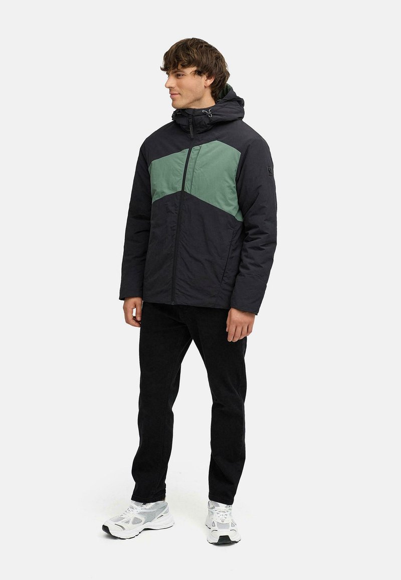 Svart och grön färgblockerad pufferjacka med dragkedja, huva och texturerad tyg, kombinerad med svarta byxor och vita sneakers.
