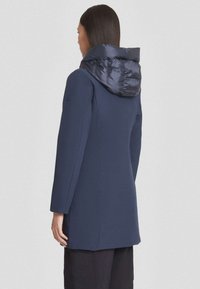 Manteau marine avec une capuche rembourrée et brillante. Présente des manches longues et une silhouette ajustée, fabriqué à partir d'un tissu lisse avec un minimum de détails.