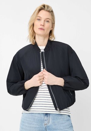 Blonde Frau trägt eine schwarze Bomberjacke über einem weiß-navy gestreiften Shirt und hellblauen Jeans und zieht den Reißverschluss der Jacke hoch.