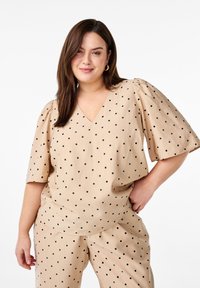 Beige Oberteil mit V-Ausschnitt und kurzen Puffärmeln, mit schwarzen Punkten. Kombiniert mit passenden beigen Hosen.