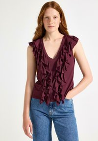 Kvinna med långt röd hår iförd ärmlös mörk lila blus med volanger och blå jeans, stående med ena handen i fickan framför en vit bakgrund.