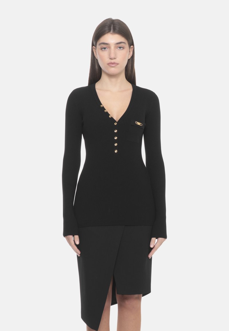 Schwarzes, geripptes Langarmoberteil mit tiefem V-Ausschnitt, goldenen Knöpfen und einer kleinen Tasche. Kombiniert mit einem schwarzen Wickelrock mit einem seitlichen Schlitz.