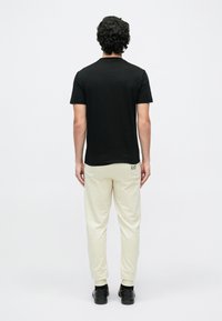 Férfi hátulról nézve, fekete rövid ujjú inget, krémszínű melegítőnadrágot, melyen apró logó van a hátsó zsebén, és fekete cipőt visel.