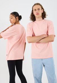 Rožinė trumpomis rankovėmis medvilnė t-shirt, turinti laisvą kirpimą. Pavaizduota priekinė ir galinė modelio, dėvinčio mėlynus džinsus, pusės.
