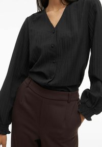 Melna blūze ar garām piedurknēm, V veida izgriezumu, vertikālas svītras un pogu aizdari. Raksturota ar savāktām manšetēm un mīkstu tekstūru. Kombinēta ar brūnām biksēm.