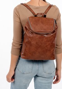 Person som bär ljusblåjeans och en beige topp med en brun läderryggsäck med ett överlock och synlig dragkedja på baksidan.