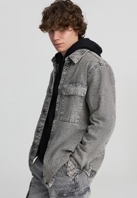 Szara koszula dżinsowa z zapinanymi kieszeniami na piersi, długimi rękawami i luźnym krojem, warstwowa na czarnym sweatshirt z kapturem.