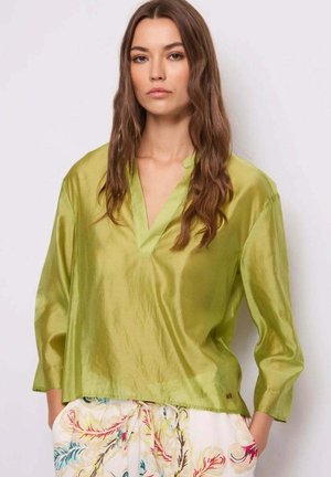 Donna con capelli castani lunghi che indossa una blusa verde lucida e ampia con scollo a V e pantaloni chiari con motivo floreale, in piedi di fronte a una parete liscia.