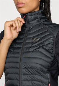 Černá izolační vesta s prošívanou texturou, vysokým límcem a růžovými zipovými detaily. Obsahuje malé logo v oblasti hrudníku.