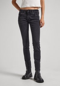 Jeans negros ajustados de denim, con cinco bolsillos, mínimo desvanecimiento y un corte slim. Combinados con botas negras de tobillo sobre un fondo claro.