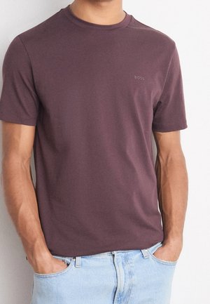 Mann trägt ein eng anliegendes dunkel mauvefarbenes Kurzarm-T-Shirt mit dem "BOSS"-Logo auf der Brust und hellblaue Jeans, Hände in den Taschen.