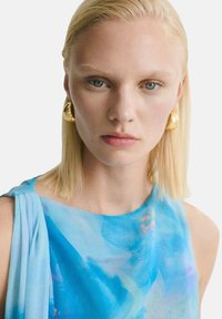 Blå blommig topp i akvarell med mjukt tyg, med vid halsringning och draperad axeldetalj. Modellen bär guldiga hoopörhängen.