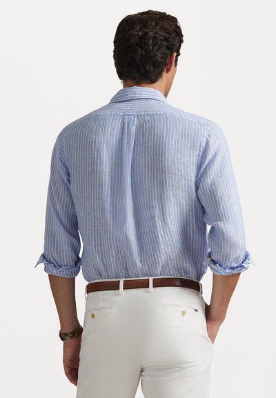 Gaiši zila un balta svītrainā krekls ar klasisku apkakli, sarullētām piedurknēm un brīvu piegriezumu, kombinēts ar baltām biksēm un brūnu jostu.