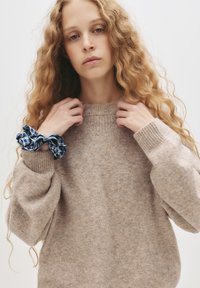 Beżowy sweter dzianinowy z prążkowanym dekoltem i mankietami, z luźnymi rękawami. Na nadgarstku noszona jest niebieska scrunchie w kratę.