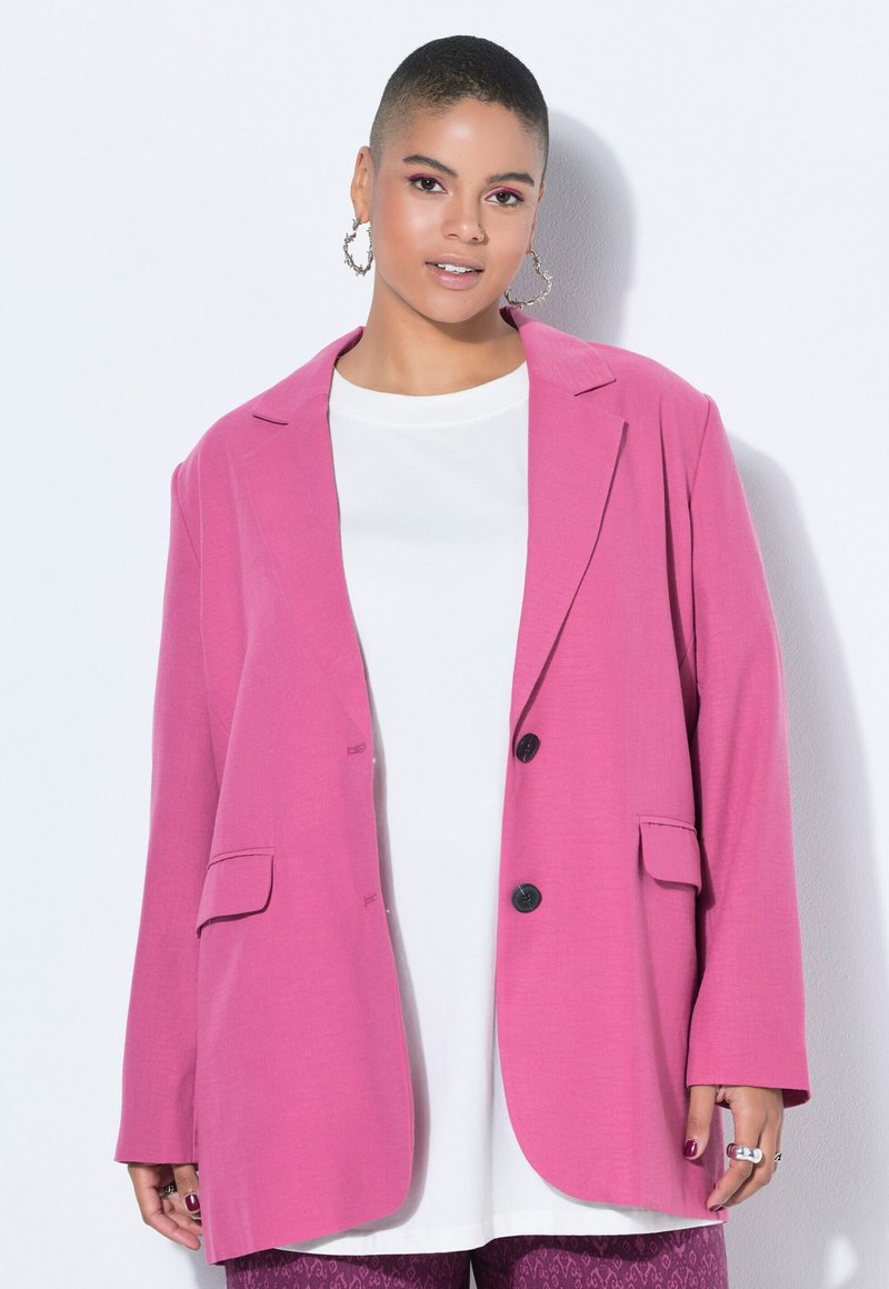 Jeune personne aux cheveux courts portant des boucles d'oreilles créoles, un blazer rose vif sur un haut blanc et un pantalon violet à motifs, debout sur fond blanc.
