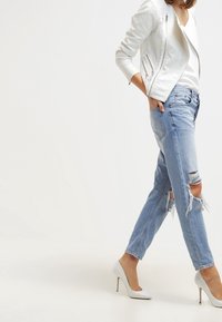 Vit perforerad skinnjacka, vit topp, urtvättade ljusblå jeans och spetsiga vita högklackade skor, visad i en dynamisk gångposition.
