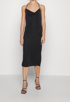 Kvinde iført en sort satin midi kjole med tynde halterstropper og sorte hæl-sandaler med remme, der står mod en ensartet lys baggrund.