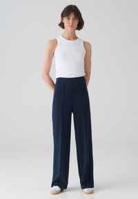 Marineblauwe hoge taille broek met wijde pijpen, plooien en een zachte textuur. Gekleed met een aansluitend wit tanktop en casual witte sneakers.