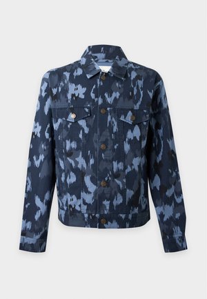 Homme portant une veste bleue à motifs sur un t-shirt blanc, un pantalon bleu et des baskets blanches, debout avec une main dans la poche.
