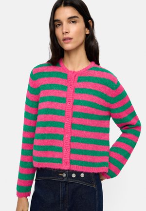 Kobieta ubrana w zapinany na guziki, puszysty kardigan z poziomymi różowymi i zielonymi paskami, w połączeniu z ciemnymi dżinsami z wysokim stanem.