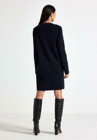 Abito lavorato a maglia blu navy con una vestibilità morbida, caratterizzato da maniche lunghe e dettagli con bottoni dorati, abbinato a stivali neri con tacco al ginocchio.