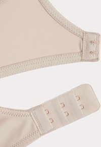 Soutien-gorge beige avec un tissu lisse, fermeture à crochets et œillets réglable, et bordure contrastante plus claire. Il présente un design sans coutures et une texture douce.