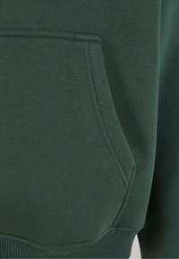 Dark green sweatshirt fabric with a large, angled pocket. Smooth texture and ribbed hem detail. Stitching is visible along the pocket edge.