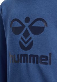 Niebieski bawełnowy sweatshirt z teksturowanym, czarnym logo w abstrakcyjnych kształtach oraz nazwą marki "hummel" w wyrazistym kroju pisma.