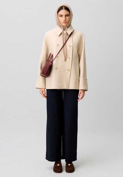 Donna che indossa un cappotto beige doppiopetto, un fazzoletto beige in testa, pantaloni neri a gamba larga, mocassini marroni e una piccola borsa a tracolla color bordeaux.