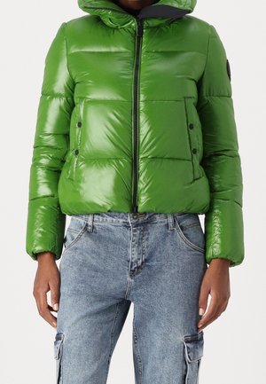 Femme portant une doudoune verte brillante avec des poches avant et un jean cargo en denim bleu.