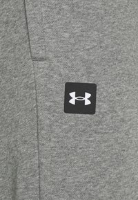 Grauer Sweatshirtstoff mit einer weichen, strukturierten Oberfläche. Verfügt über ein schwarzes quadratisches Patch mit einem weißen Under Armour-Logo.