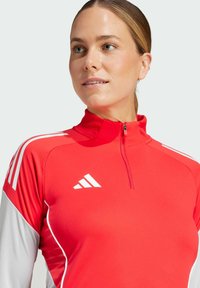 Rød sports top med halv-zip halsudskæring, med hvide accenter og tre striber på skuldrene; lavet af glat, strækbart materiale.
