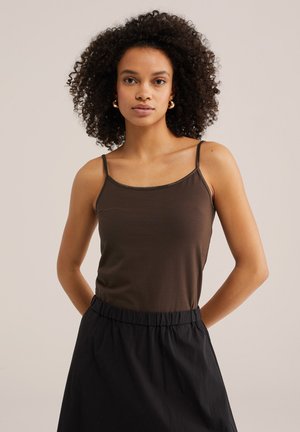 Mujer con cabello rizado, vestida con un top marrón de tirantes finos y pantalones negros de talle alto, de pie contra un fondo claro y sencillo.