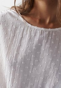Blusa bianca ricamata caratterizzata da un tessuto testurizzato con motivi a freccia in rilievo, scollatura rotonda e maniche corte.