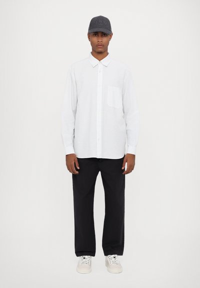 Camisa blanca de botones con un bolsillo en el pecho, combinada con pantalones negros y zapatillas blancas, complementada con una gorra gris oscuro con logotipo.