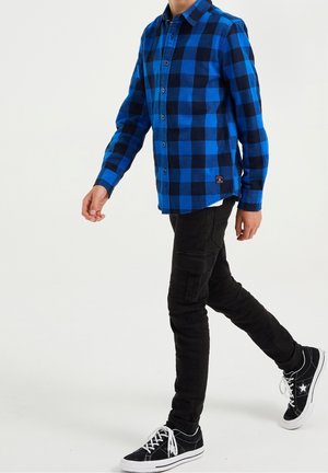 Blå og sort ternet skjorte med knapper, bomuldsstof, lange ærmer, kombineret med sorte slim-fit bukser og sorte sneakers med hvide detaljer.