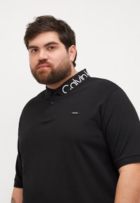 Μαύρο πόλο μπλουζάκι με κοντά μανίκια, με υφή υφάσματος, που διαθέτει γιακά με αντίθεση μαύρου και λευκού λογότυπου και μικρό λογότυπο στο στήθος.