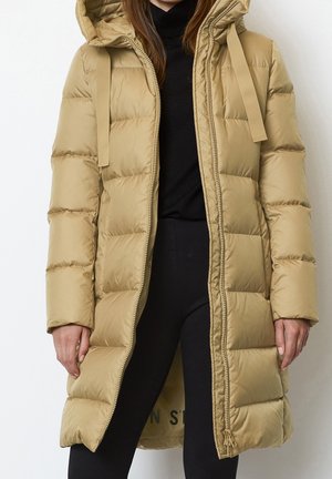 Femme portant un long manteau matelassé beige à capuche, ouvert sur un col roulé noir et un pantalon noir, posant devant un fond blanc.