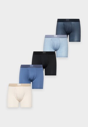 Un homme torse nu présente un boxer bleu. Il se tient debout, les mains derrière le dos, et regarde sur le côté devant un fond blanc uni.
