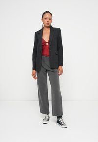 Jeune femme portant un blazer à carreaux noirs, un haut en dentelle rouge, un pantalon large gris et des baskets montantes noires, se tenant contre un fond blanc.