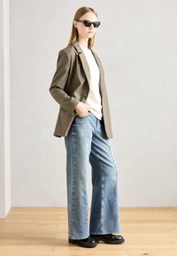 Ελαιολαδί σακάκι πάνω από κρεμ μπλούζα σε συνδυασμό με φαρδιά μπλε τζιν και μαύρες πλατφόρμες· το μοντέλο φορά γωνιακά μαύρα γυαλιά ηλίου.