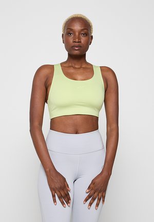 Femme aux cheveux blonds courts portant un brassard de sport vert clair et un legging gris clair taille haute, debout les mains détendues devant elle.
